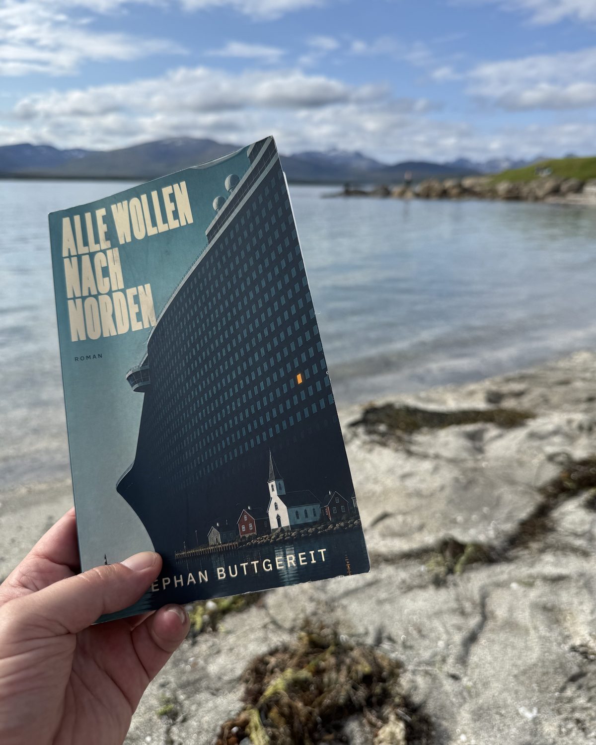 Das Taschenbuch Alle wollen nach Norden, in einer Hand gehalten am Stadtstrand von Tromsø, dahinter Fjord und Berge